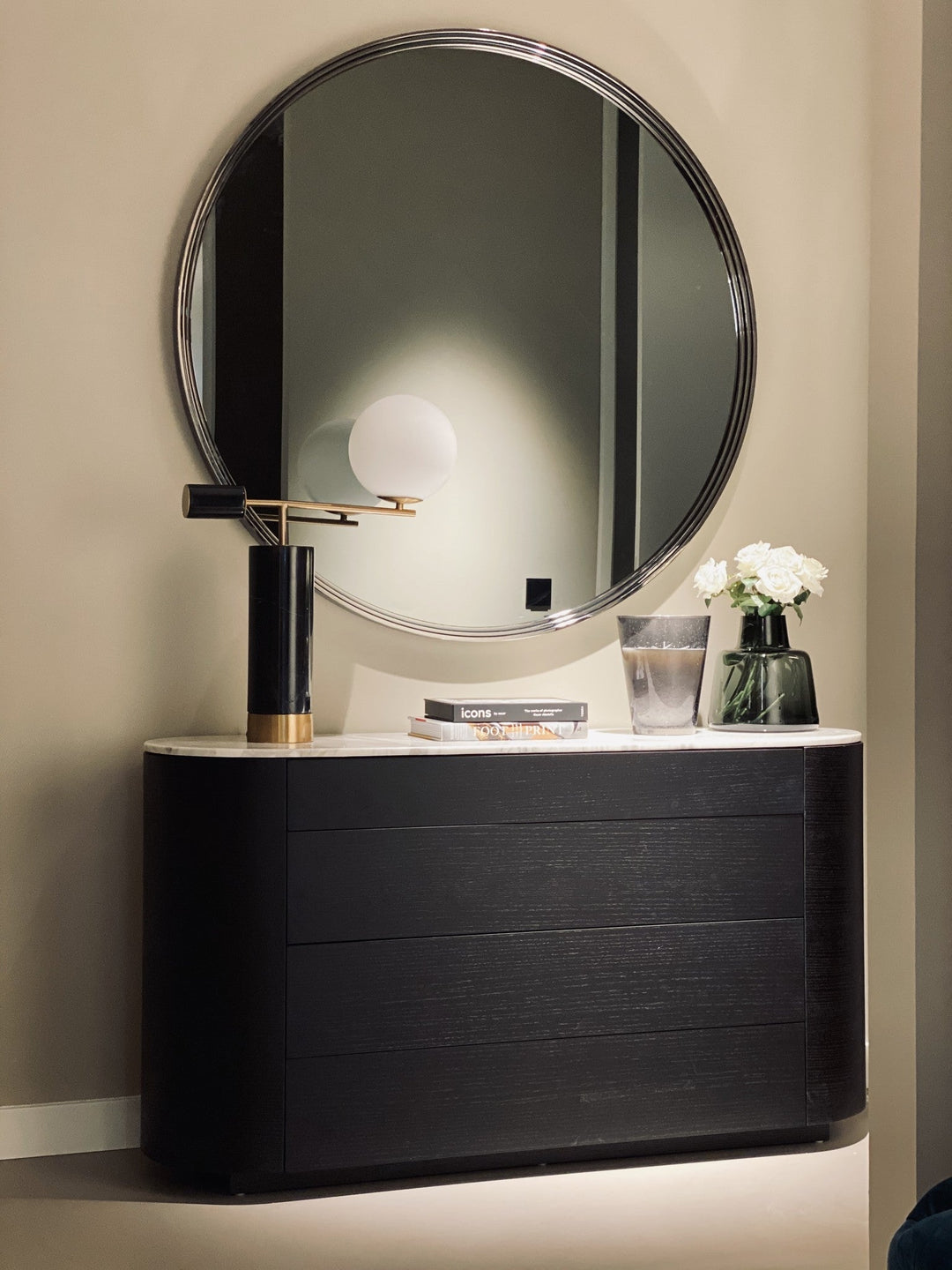 Dressoir Aldo in eiken Charcoal met een licht marmeren blad, gestyled met een tafellamp en bloemen onder een grote ronde spiegel.