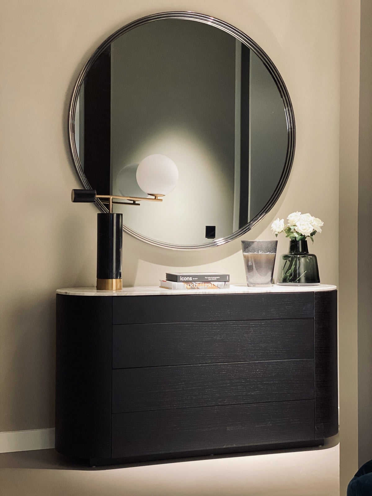 Dressoir Aldo in eiken Charcoal met een licht marmeren blad, gestyled met een tafellamp en bloemen onder een grote ronde spiegel.