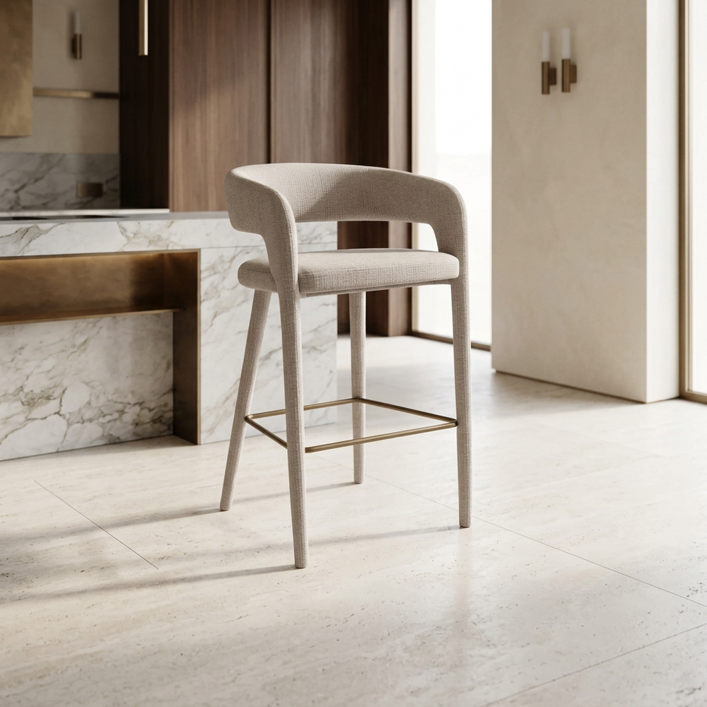 Barstoel Valeria met licht gestoffeerde bekleding en bronzen voetsteun bij een marmeren keukenbar in een warm interieur.